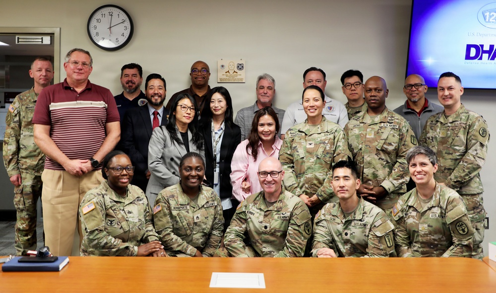 A group photo of approximately 20 individuals, a mix of U.S. Army military personnel in camouflage uniforms and civilians in business casual attire, posed in what appears to be a conference or briefing room. The setting includes a wall clock, a commemorative plaque, and a monitor displaying a DHA (Defense Health Agency) logo. The group is arranged in three rows with front row seated, suggesting a formal organizational or unit photo.