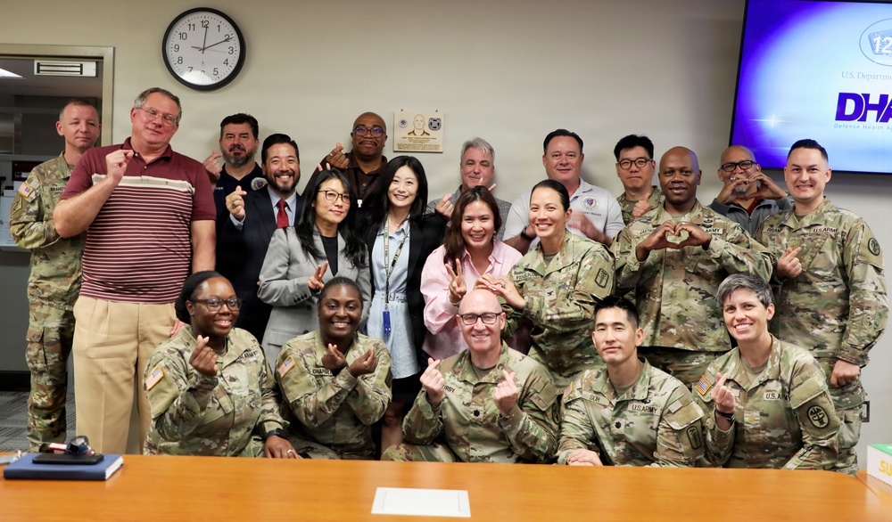 A group photo of approximately 20 individuals, predominantly U.S. Army personnel in camouflage uniforms, along with several civilians, posing in what appears to be a conference or office room. Most participants are making finger-heart or similar hand gestures toward the camera. A DHA (Defense Health Agency) logo is visible on a screen in the background.