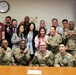 A group photo of approximately 20 individuals, predominantly U.S. Army personnel in camouflage uniforms, along with several civilians, posing in what appears to be a conference or office room. Most participants are making finger-heart or similar hand gestures toward the camera. A DHA (Defense Health Agency) logo is visible on a screen in the background.