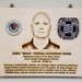 A wooden memorial plaque dedicated to James 'Brian' Connor, Chief of Facilities Management Division, mounted on a wall with two metal hanging hooks. The plaque features a laser-engraved portrait of the honoree along with two military/agency medallions. It commemorates his service dates of 4 Nov 69 – 10 Dec 25 and his contributions to military medical infrastructure.
