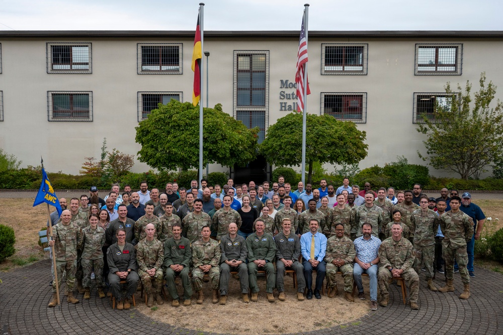 A large group photo of approximately 80-100 military personnel and civilians posing in front of a building identified as 'Moo... Sut... Hall' (partially obscured). The group is arranged in multiple rows with seated individuals in the front and standing rows behind them. Two flags are visible — the German flag and the United States flag — indicating this is likely a U.S. military installation in Germany.
