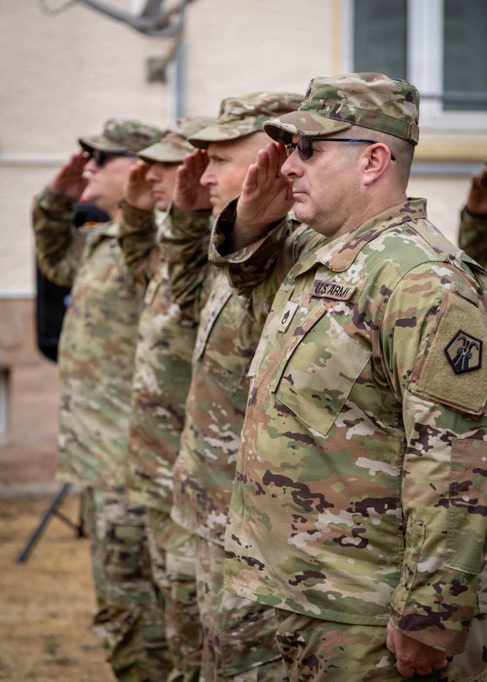 A group of U.S. Army soldiers in Operational Camouflage Pattern (OCP) uniforms are standing in a line and rendering a salute during what appears to be a formal military ceremony. The foreground soldier displays a 'U.S. ARMY' tape and a unit patch on the left sleeve. The setting appears to be an outdoor military installation.