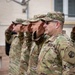 A group of U.S. Army soldiers in Operational Camouflage Pattern (OCP) uniforms are standing in a line and rendering a salute during what appears to be a formal military ceremony. The foreground soldier displays a 'U.S. ARMY' tape and a unit patch on the left sleeve. The setting appears to be an outdoor military installation.