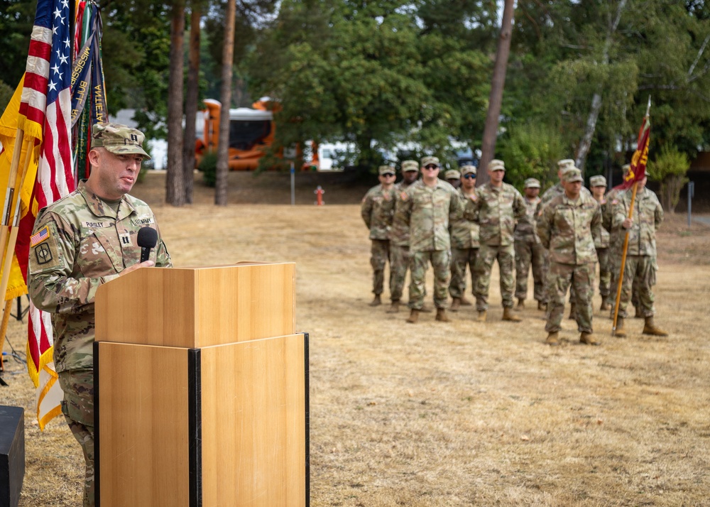 A U.S. Army officer, identifiable by captain's rank insignia, speaks at a podium during what appears to be a military ceremony outdoors. An American flag and unit guidons are displayed beside the podium, while a formation of soldiers in OCP (Operational Camouflage Pattern) uniforms stands at ease in the background. The setting appears to be a military installation, likely in Europe based on the environment.