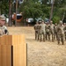 A U.S. Army officer, identifiable by captain's rank insignia, speaks at a podium during what appears to be a military ceremony outdoors. An American flag and unit guidons are displayed beside the podium, while a formation of soldiers in OCP (Operational Camouflage Pattern) uniforms stands at ease in the background. The setting appears to be a military installation, likely in Europe based on the environment.