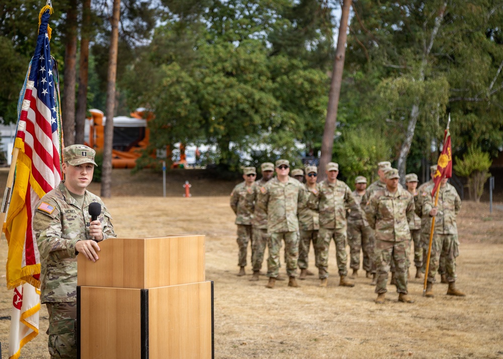 A U.S. Army officer is speaking at a podium during what appears to be a formal military ceremony held outdoors. The speaker stands beside an American flag and another military flag, addressing a formation of soldiers standing at ease in the background. The setting appears to be a military installation or training area with trees and equipment visible in the background.