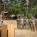 A U.S. Army officer is speaking at a podium during what appears to be a formal military ceremony held outdoors. The speaker stands beside an American flag and another military flag, addressing a formation of soldiers standing at ease in the background. The setting appears to be a military installation or training area with trees and equipment visible in the background.
