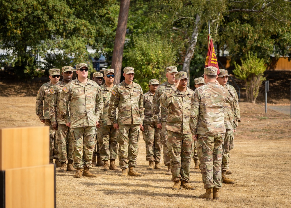 A group of U.S. Army soldiers in Operational Camouflage Pattern (OCP) uniforms are standing in formation on a dry grass field. An officer or senior NCO appears to be addressing or inspecting the formation, with a red unit guidon flag visible in the background. The setting appears to be an outdoor military ceremony or formation event.