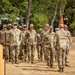 A group of U.S. Army soldiers in Operational Camouflage Pattern (OCP) uniforms are standing in formation on a dry grass field. An officer or senior NCO appears to be addressing or inspecting the formation, with a red unit guidon flag visible in the background. The setting appears to be an outdoor military ceremony or formation event.
