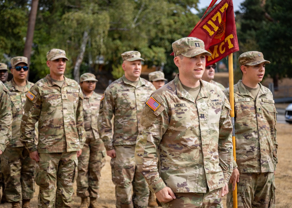 A group of U.S. Army soldiers in MultiCam/OCP camouflage uniforms are assembled outdoors, likely during a military ceremony or formation. The central figure, identified by name tape as 'MCNEIL,' holds a red unit guidon bearing the designation '117 MCT.' Soldiers in the background include individuals identified as 'PEREZ' and 'STEELE,' with various ranks visible including a Captain's rank insignia on MCNEIL.