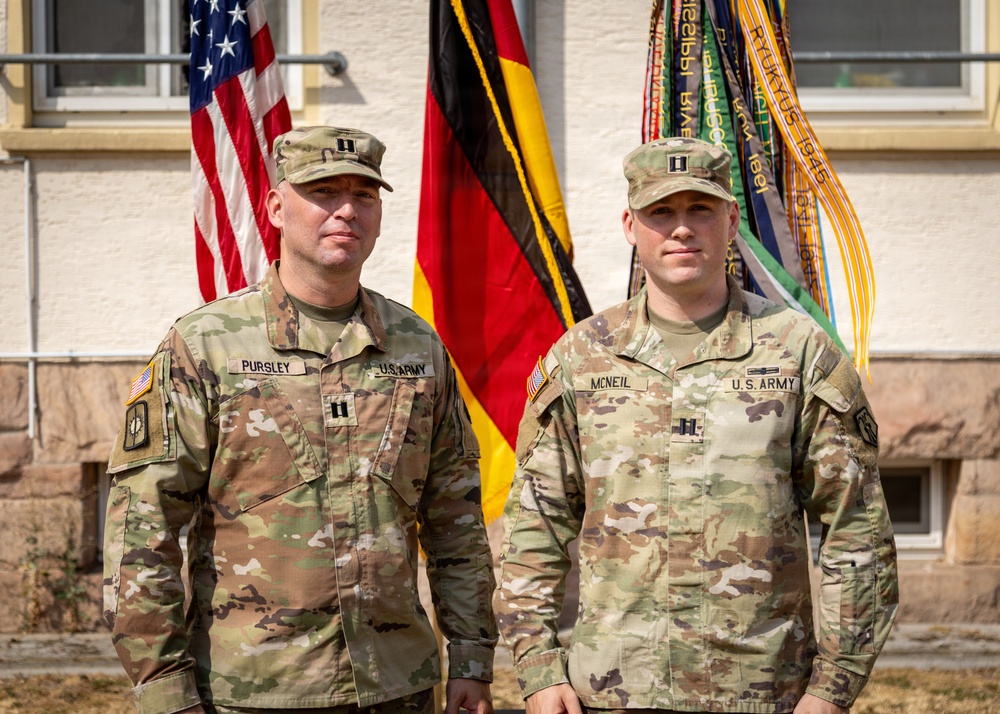 Two U.S. Army Captains in OCP (Operational Camouflage Pattern) uniforms pose together in front of an American flag, a German flag, and a unit guidon with campaign streamers. Their name tapes identify them as PURSLEY (left) and MCNEIL (right), both wearing Captain rank insignia (two silver bars) on their caps and chest. The setting appears to be an outdoor military ceremony or official photo at a U.S. installation in Germany.