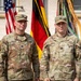 Two U.S. Army Captains in OCP (Operational Camouflage Pattern) uniforms pose together in front of an American flag, a German flag, and a unit guidon with campaign streamers. Their name tapes identify them as PURSLEY (left) and MCNEIL (right), both wearing Captain rank insignia (two silver bars) on their caps and chest. The setting appears to be an outdoor military ceremony or official photo at a U.S. installation in Germany.