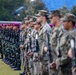 A formal military parade or ceremony featuring two distinct groups of soldiers standing in formation on a grass field. One group wears distinctive magenta/purple berets with green camouflage uniforms, while the other group wears digital camouflage uniforms with patrol caps, suggesting a joint multinational military exercise or ceremony. All personnel are armed with rifles and standing at attention.