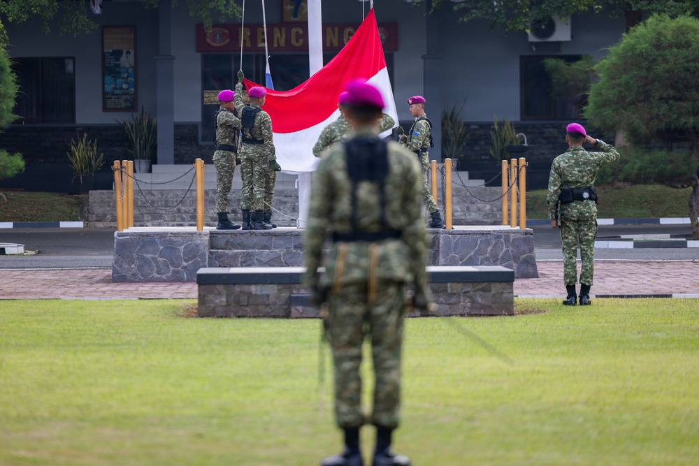 A formal military flag-raising ceremony is taking place at what appears to be an Indonesian military installation. Several soldiers wearing camouflage uniforms and distinctive purple/magenta berets are participating, with two soldiers on a raised stone platform hoisting a red-and-white Indonesian national flag (Merah Putih). Other personnel stand at attention or salute in the foreground and to the side.