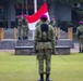 A formal military flag-raising ceremony is taking place at what appears to be an Indonesian military installation. Several soldiers wearing camouflage uniforms and distinctive purple/magenta berets are participating, with two soldiers on a raised stone platform hoisting a red-and-white Indonesian national flag (Merah Putih). Other personnel stand at attention or salute in the foreground and to the side.