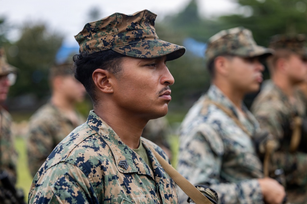 A group of military personnel in digital camouflage uniforms are standing in formation outdoors. The foreground subject, wearing MARPAT (Marine Pattern) camouflage and a rank insignia consistent with a Sergeant chevron, is in sharp focus while others are blurred in the background. The setting appears to be an outdoor military training or ceremonial event.