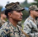 A group of military personnel in digital camouflage uniforms are standing in formation outdoors. The foreground subject, wearing MARPAT (Marine Pattern) camouflage and a rank insignia consistent with a Sergeant chevron, is in sharp focus while others are blurred in the background. The setting appears to be an outdoor military training or ceremonial event.