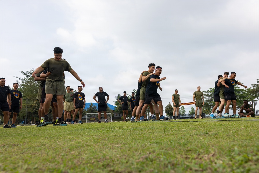 A group of military or paramilitary personnel are engaged in outdoor physical training exercises on a grass field. Participants are wearing mixed uniforms of olive/khaki and black athletic wear, some with headbands, suggesting a joint or combined training exercise. The setting appears to be a military or sports facility in a tropical region based on the vegetation and architecture visible in the background.