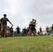 A group of military or paramilitary personnel are engaged in outdoor physical training exercises on a grass field. Participants are wearing mixed uniforms of olive/khaki and black athletic wear, some with headbands, suggesting a joint or combined training exercise. The setting appears to be a military or sports facility in a tropical region based on the vegetation and architecture visible in the background.