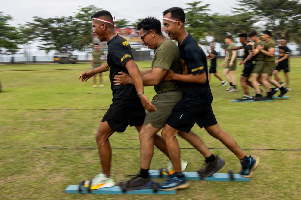 A group of individuals are participating in a team coordination exercise on a grass field, where participants appear to be moving together on shared platforms or boards. Two individuals in black uniforms with red-and-white headbands are flanking a person in an olive green shirt and glasses, all moving in unison. In the background, additional pairs or groups are performing the same activity, suggesting a structured team-building or military training event.