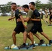 A group of individuals are participating in a team coordination exercise on a grass field, where participants appear to be moving together on shared platforms or boards. Two individuals in black uniforms with red-and-white headbands are flanking a person in an olive green shirt and glasses, all moving in unison. In the background, additional pairs or groups are performing the same activity, suggesting a structured team-building or military training event.