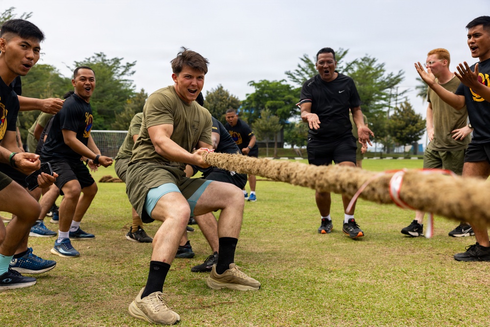 A group of military personnel are engaged in a tug-of-war competition on a grass field. The participants appear to represent two different units or nationalities, with some wearing olive/tan military PT gear and others wearing black t-shirts with unit insignia. The atmosphere is energetic and competitive, with participants visibly straining and cheering.