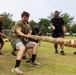 A group of military personnel are engaged in a tug-of-war competition on a grass field. The participants appear to represent two different units or nationalities, with some wearing olive/tan military PT gear and others wearing black t-shirts with unit insignia. The atmosphere is energetic and competitive, with participants visibly straining and cheering.