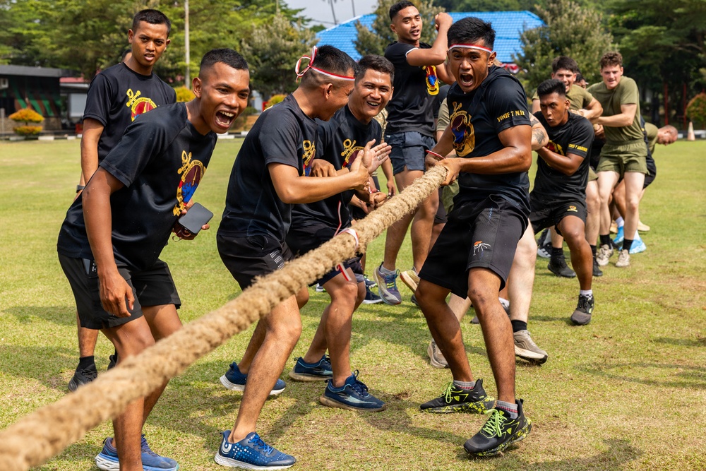 A group of military personnel are engaged in a tug-of-war competition on a grass field. The majority of participants wear matching black t-shirts with a military unit insignia, while a smaller group in the background wear olive/green military-style shirts, suggesting an inter-unit or multinational friendly competition. The atmosphere is energetic and celebratory, with participants visibly straining and cheering.