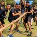 A group of military personnel are engaged in a tug-of-war competition on a grass field. The majority of participants wear matching black t-shirts with a military unit insignia, while a smaller group in the background wear olive/green military-style shirts, suggesting an inter-unit or multinational friendly competition. The atmosphere is energetic and celebratory, with participants visibly straining and cheering.