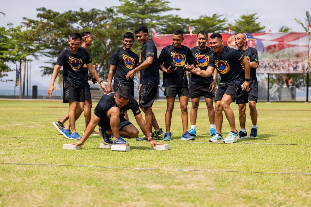 A group of men wearing matching black uniforms with an emblem are participating in an outdoor team activity on a grass field. One participant is crouched low to the ground, hands on two flat stone or concrete blocks, while teammates cheer and laugh around him. The setting appears to be a military or institutional sports event, with banners and a crowd visible in the background.