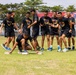 A group of men wearing matching black uniforms with an emblem are participating in an outdoor team activity on a grass field. One participant is crouched low to the ground, hands on two flat stone or concrete blocks, while teammates cheer and laugh around him. The setting appears to be a military or institutional sports event, with banners and a crowd visible in the background.
