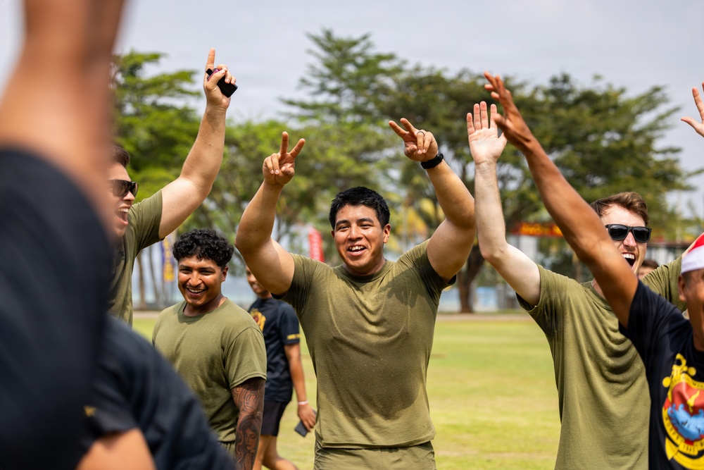 A group of young men wearing olive green t-shirts are celebrating outdoors on a grass field, raising their arms and making hand gestures with big smiles. The setting appears to be a military or organized sporting event, with participants showing clear signs of victory or excitement. The atmosphere is energetic and celebratory.