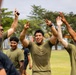 A group of young men wearing olive green t-shirts are celebrating outdoors on a grass field, raising their arms and making hand gestures with big smiles. The setting appears to be a military or organized sporting event, with participants showing clear signs of victory or excitement. The atmosphere is energetic and celebratory.