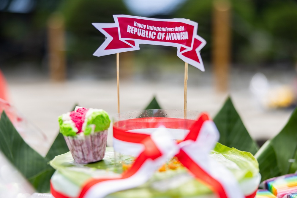 The image shows a festive display celebrating Indonesian Independence Day, featuring a decorative banner reading 'Happy Independence Day 61st Republic of Indonesia' mounted on wooden skewers. The arrangement includes traditional Indonesian elements such as banana leaves, a colorful cupcake, and red-and-white decorations matching the Indonesian national flag colors. The setup appears to be part of an outdoor celebration or ceremonial display.