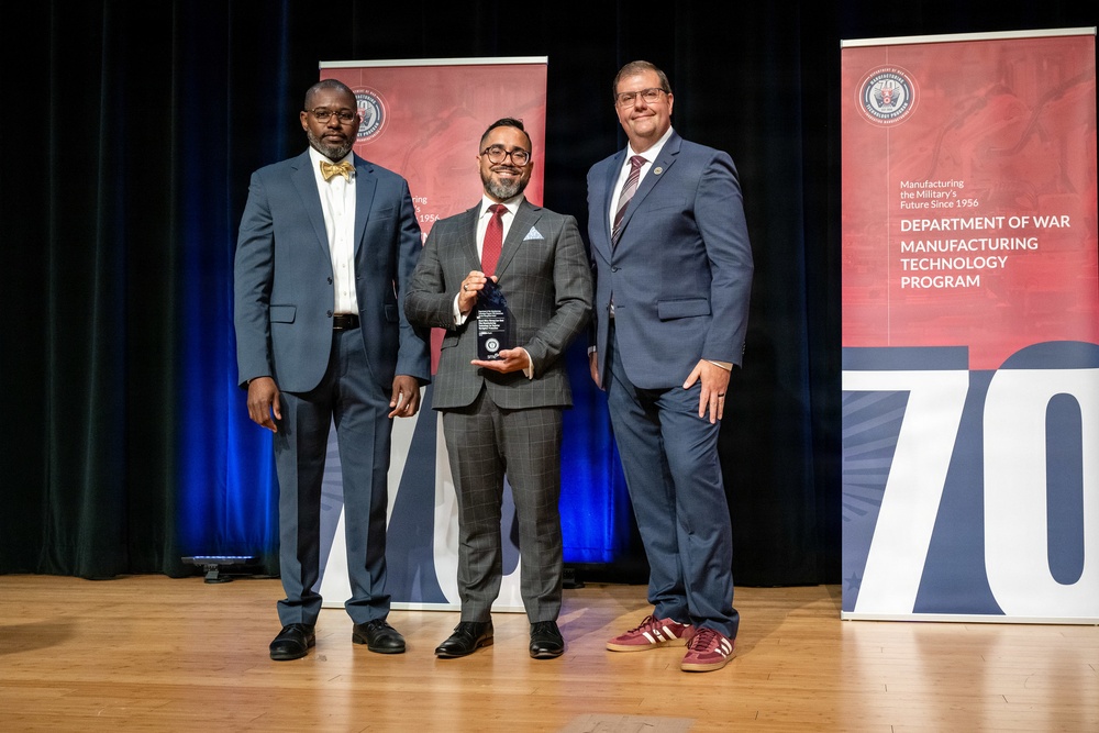 Three men in business attire are posed on a stage for a formal award presentation. The central figure is holding a crystal or glass trophy award. The backdrop includes banners for the Department of War Manufacturing Technology Program, celebrating its 70th anniversary since 1956.