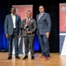 Three men in business attire are posed on a stage for a formal award presentation. The central figure is holding a crystal or glass trophy award. The backdrop includes banners for the Department of War Manufacturing Technology Program, celebrating its 70th anniversary since 1956.