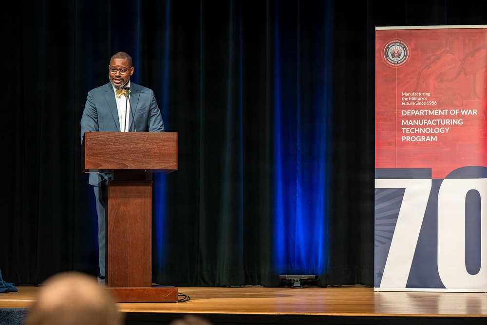 A man in a gray suit and yellow bow tie is speaking at a wooden podium on a stage. Behind him is a banner for the 'Department of War Manufacturing Technology Program,' noting it has been 'Manufacturing the Military's Future Since 1956.' The event appears to be a formal ceremony or conference, likely celebrating a milestone anniversary (the number '70' is prominently displayed on the lower banner).
