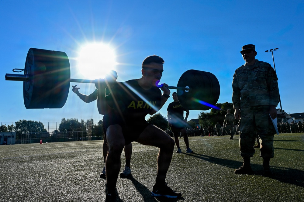 U.S. Army soldiers are performing barbell squats on an outdoor athletic field during what appears to be a physical fitness test or training event. A uniformed observer in camouflage pattern (ACU/OCP) stands nearby, likely serving as a grader or supervisor holding a clipboard. The image is dramatically backlit by a bright sun, creating strong silhouettes and lens flare effects.