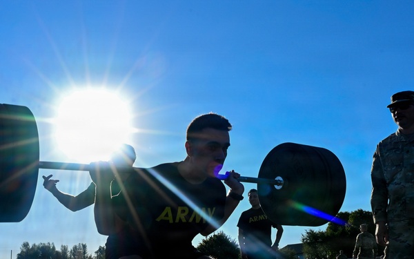 U.S. Army soldiers are performing barbell squats on an outdoor athletic field during what appears to be a physical fitness test or training event. A uniformed observer in camouflage pattern (ACU/OCP) stands nearby, likely serving as a grader or supervisor holding a clipboard. The image is dramatically backlit by a bright sun, creating strong silhouettes and lens flare effects.