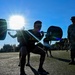 U.S. Army soldiers are performing barbell squats on an outdoor athletic field during what appears to be a physical fitness test or training event. A uniformed observer in camouflage pattern (ACU/OCP) stands nearby, likely serving as a grader or supervisor holding a clipboard. The image is dramatically backlit by a bright sun, creating strong silhouettes and lens flare effects.