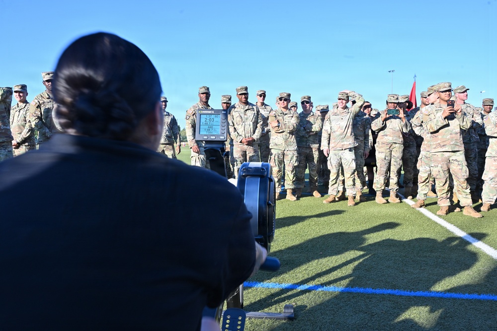 A person in civilian clothing is using a rowing machine (ergometer) on a turf field while a large group of uniformed military personnel in OCP (Operational Camouflage Pattern) uniforms observe. The setting appears to be an outdoor athletic field, likely during a fitness demonstration or physical training event. A red guidon flag is visible among the formation of soldiers.