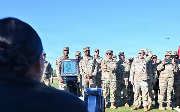 A person in civilian clothing is using a rowing machine (ergometer) on a turf field while a large group of uniformed military personnel in OCP (Operational Camouflage Pattern) uniforms observe. The setting appears to be an outdoor athletic field, likely during a fitness demonstration or physical training event. A red guidon flag is visible among the formation of soldiers.