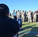 A person in civilian clothing is using a rowing machine (ergometer) on a turf field while a large group of uniformed military personnel in OCP (Operational Camouflage Pattern) uniforms observe. The setting appears to be an outdoor athletic field, likely during a fitness demonstration or physical training event. A red guidon flag is visible among the formation of soldiers.