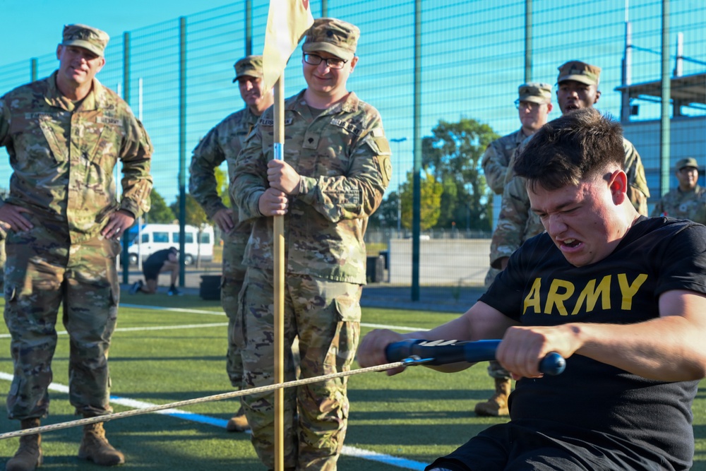 A U.S. Army soldier in PT (physical training) gear is straining intensely while pulling a rope or cable using a handle bar, likely during a fitness competition or training exercise. Several soldiers in OCP (Operational Camouflage Pattern) uniforms observe the activity, with one holding a unit guidon flag. The event appears to take place on an outdoor athletic field with a chain-link fence visible in the background.