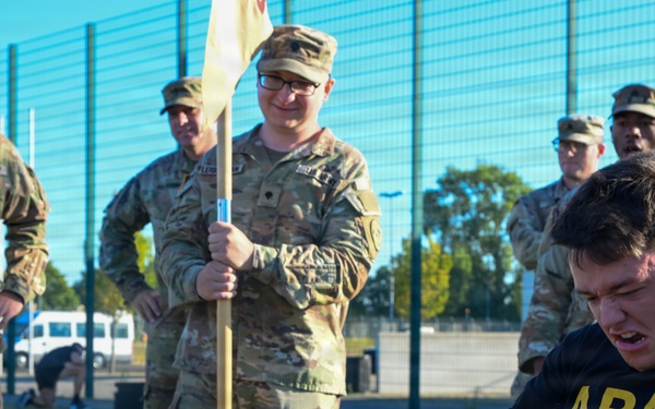 A U.S. Army soldier in PT (physical training) gear is straining intensely while pulling a rope or cable using a handle bar, likely during a fitness competition or training exercise. Several soldiers in OCP (Operational Camouflage Pattern) uniforms observe the activity, with one holding a unit guidon flag. The event appears to take place on an outdoor athletic field with a chain-link fence visible in the background.