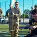 A U.S. Army soldier in PT (physical training) gear is straining intensely while pulling a rope or cable using a handle bar, likely during a fitness competition or training exercise. Several soldiers in OCP (Operational Camouflage Pattern) uniforms observe the activity, with one holding a unit guidon flag. The event appears to take place on an outdoor athletic field with a chain-link fence visible in the background.