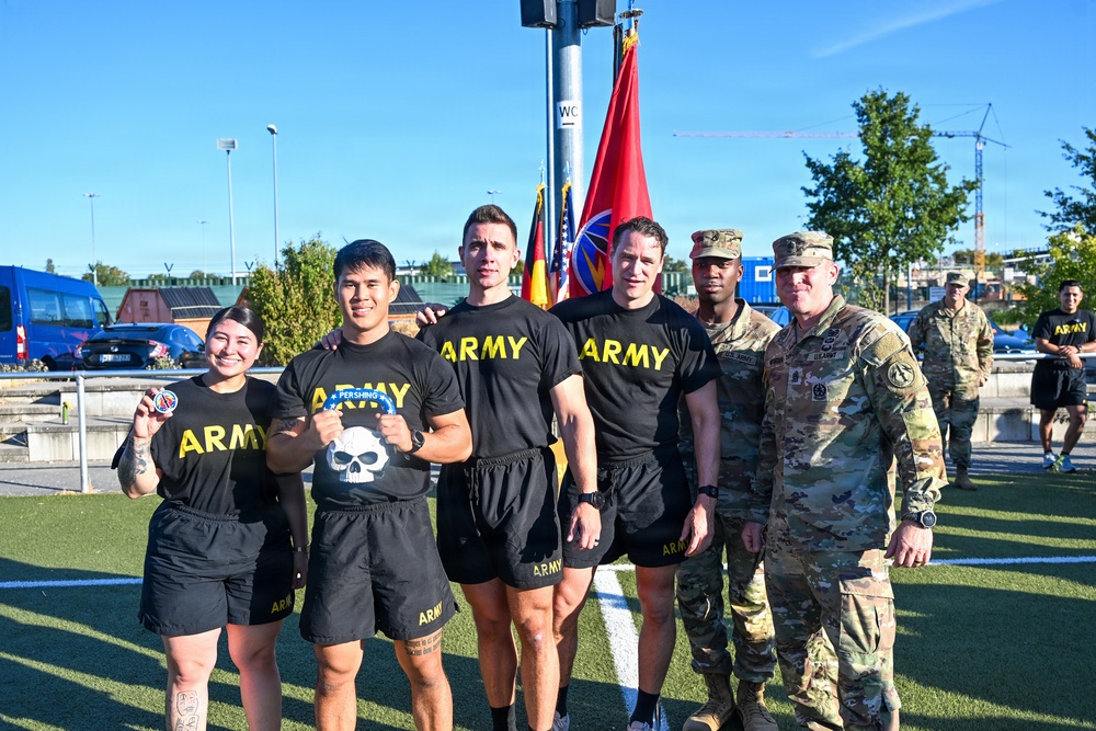 A group of six individuals pose for a photo outdoors on a green turf field, with four wearing black 'ARMY' PT (physical training) shirts and shorts, and two in military camouflage uniforms (OCP pattern). Flags including what appears to be a U.S. Army unit guidon and a German flag are visible in the background. The setting appears to be a U.S. military installation, likely in Germany based on the German flag and European-style buildings.