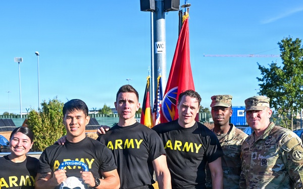 A group of six individuals pose for a photo outdoors on a green turf field, with four wearing black 'ARMY' PT (physical training) shirts and shorts, and two in military camouflage uniforms (OCP pattern). Flags including what appears to be a U.S. Army unit guidon and a German flag are visible in the background. The setting appears to be a U.S. military installation, likely in Germany based on the German flag and European-style buildings.