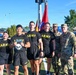 A group of six individuals pose for a photo outdoors on a green turf field, with four wearing black 'ARMY' PT (physical training) shirts and shorts, and two in military camouflage uniforms (OCP pattern). Flags including what appears to be a U.S. Army unit guidon and a German flag are visible in the background. The setting appears to be a U.S. military installation, likely in Germany based on the German flag and European-style buildings.
