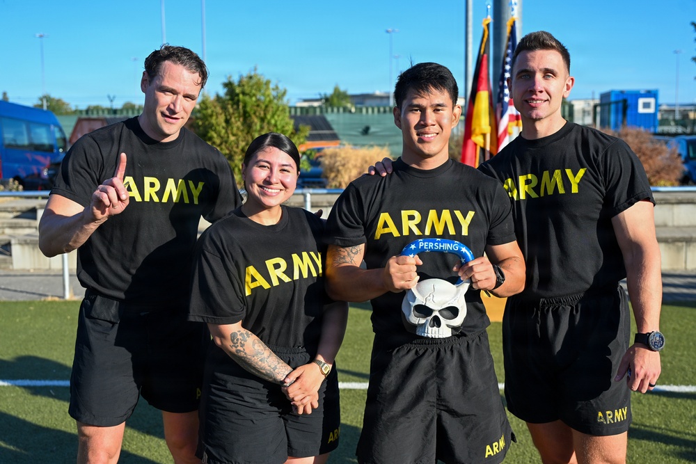 Four U.S. Army personnel pose together outdoors in matching black Army PT (Physical Training) shirts and shorts. One individual holds a skull-shaped trophy or award labeled 'PERSHING,' and another gives a thumbs-up gesture. The group appears to be celebrating a competition or event victory, with U.S. and German flags visible in the background.